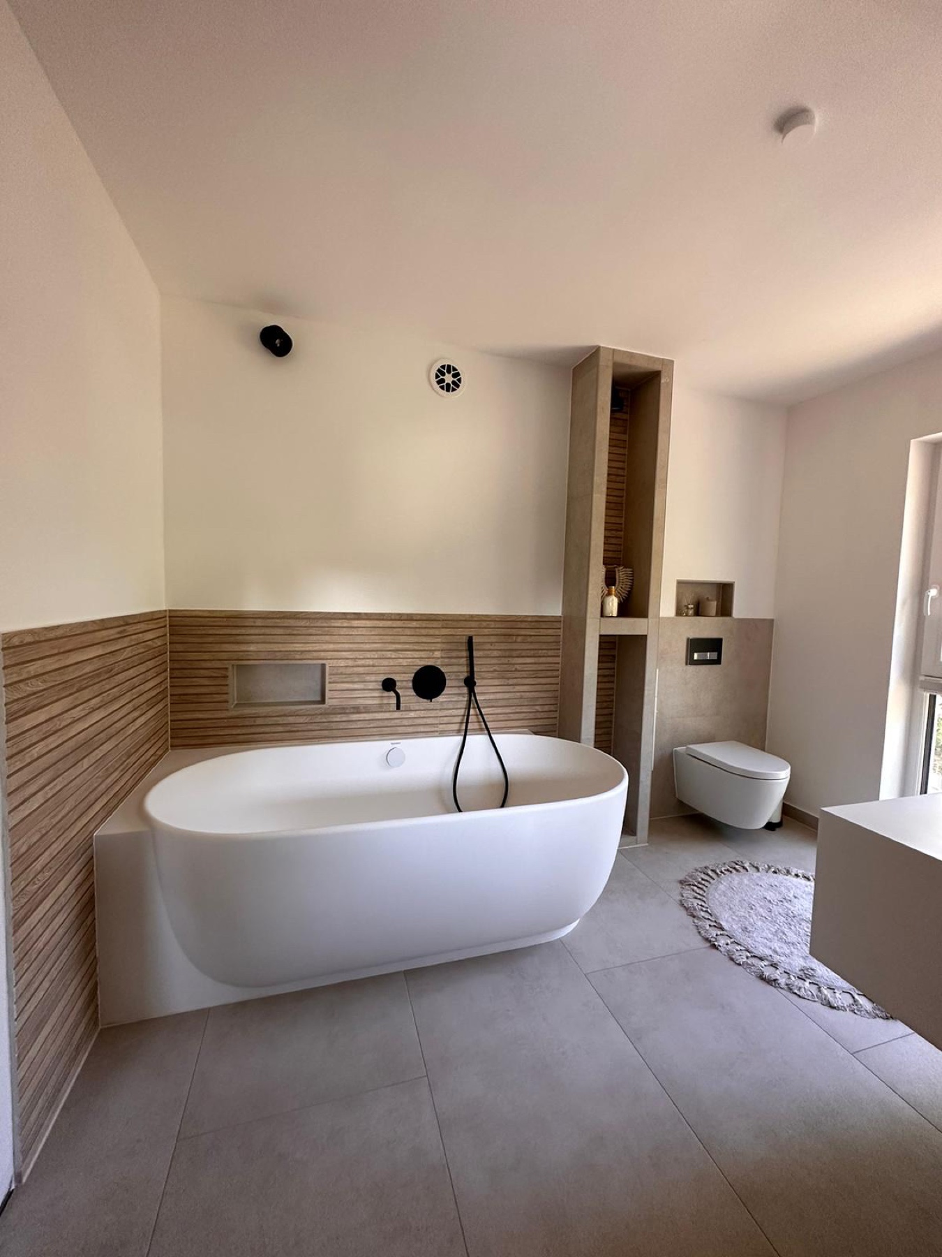 Freistehende Wanne vor warmer Wandgestaltung und grauem Fliesenboden